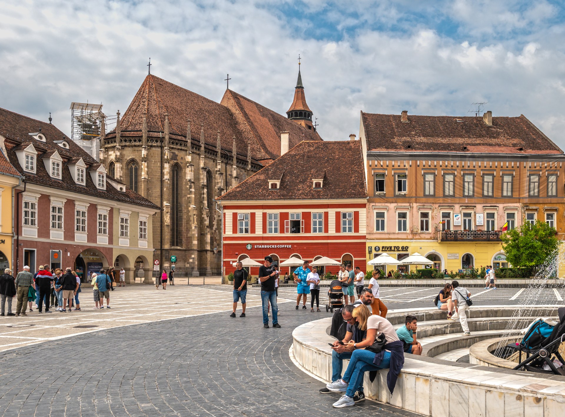Mladí ľudia oddychujú pri fontáne na Radničnom námestí v Brașove, v pozadí Čierny kostol